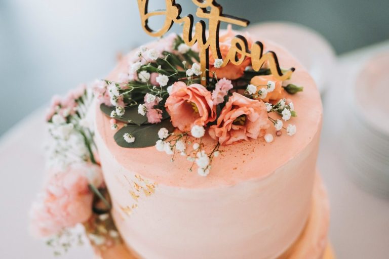 Zwei-stöckiger Torte mit rosa Frosting, Blumen und goldener Aufschrift "Britton".