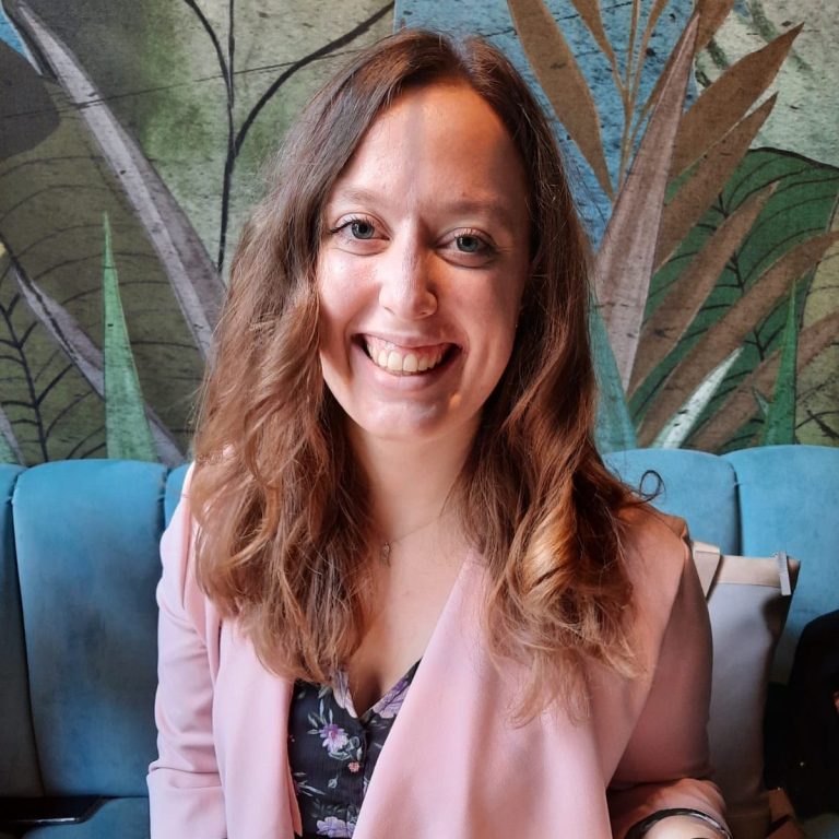 Lächelnde Frau mit langen, lockigen Haaren, in einer hellen Umgebung mit Pflanzenmotiven.