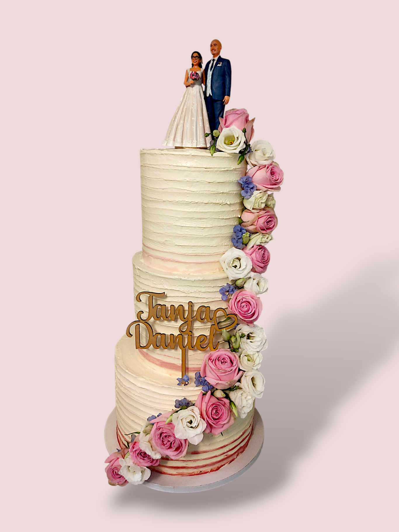 Dreistöckige Hochzeitstorte mit Blumen und dekorativem Schriftzug und einer Figur des Brautpaares oben.